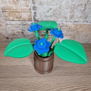 May include: A decorative, 3D-printed flower arrangement. The arrangement features bright blue flowers with green stems and leaves, set in a brown barrel-shaped pot. The leaves are large and heart-shaped.