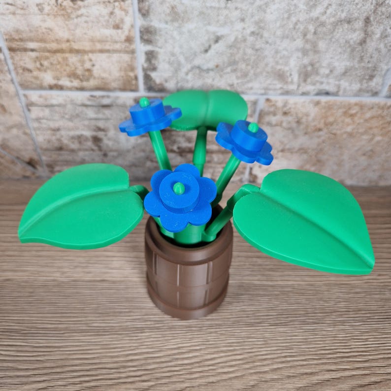 May include: A decorative, 3D-printed flower arrangement. The arrangement features bright blue flowers with green stems and leaves, set in a brown barrel-shaped pot. The leaves are large and heart-shaped.