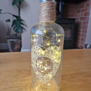 May include: A clear glass bottle with a cork top and a heart-shaped design with the word "love" inside. The bottle is wrapped with twine and contains a string of fairy lights.