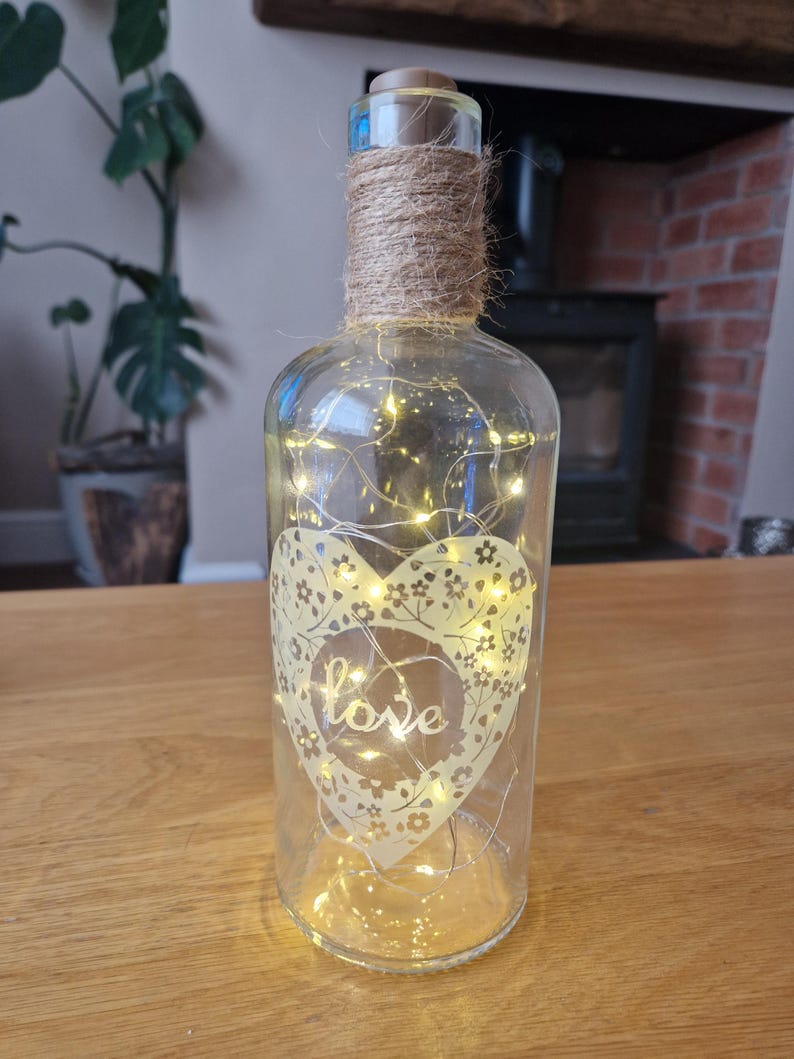 May include: A clear glass bottle with a cork top and a heart-shaped design with the word "love" inside. The bottle is wrapped with twine and contains a string of fairy lights.