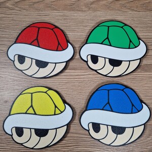 K&ouml;nnte beinhalten: Vier bunte Untersetzer in Form von Comic-Schildkr&ouml;tenpanzern. Jeder Untersetzer hat eine andere Panzerfarbe: rot, gr&uuml;n, gelb und blau. Die Panzer haben einen wei&szlig;en Rand und ein beiges Gesicht mit schwarzen Augen. Die Untersetzer liegen auf einer Holzmaserung.