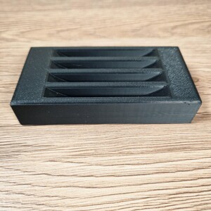 May include: A black, rectangular object with a series of angled slots. The object is made of a textured material and sits on a light brown wooden surface. The slots are evenly spaced across the top.