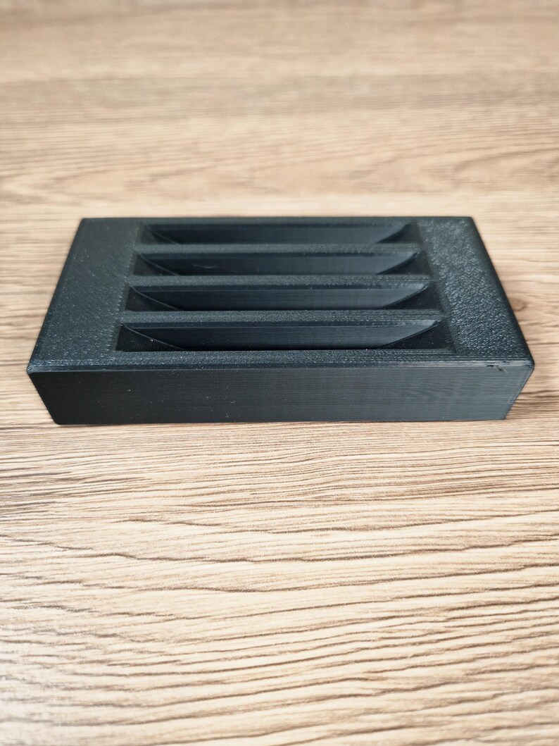 May include: A black, rectangular object with a series of angled slots. The object is made of a textured material and sits on a light brown wooden surface. The slots are evenly spaced across the top.