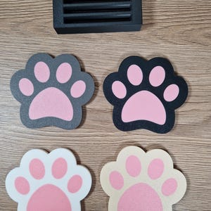 Op de afbeelding: Een zwart rechthoekig ventilatierooster en vier pootvormige onderzetters. De onderzetters zijn in verschillende kleuren: zwart met roze, grijs met roze, wit met roze en beige met roze. De items liggen op een houten oppervlak.