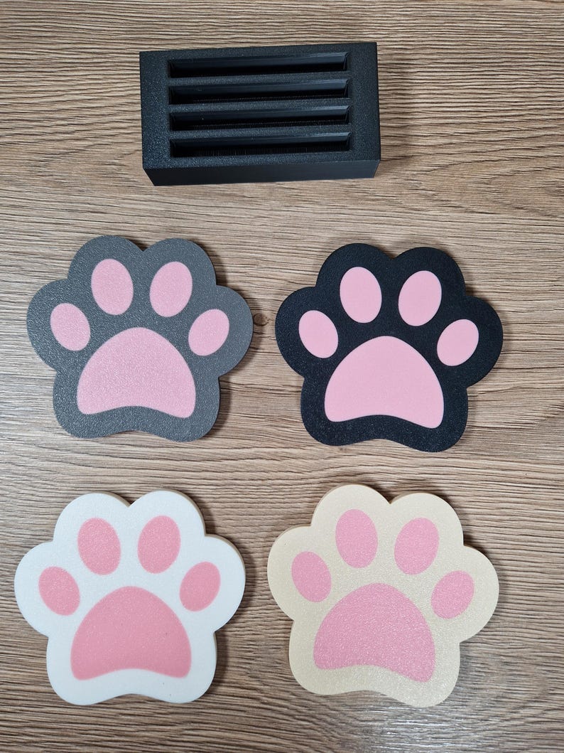 Op de afbeelding: Een zwart rechthoekig ventilatierooster en vier pootvormige onderzetters. De onderzetters zijn in verschillende kleuren: zwart met roze, grijs met roze, wit met roze en beige met roze. De items liggen op een houten oppervlak.