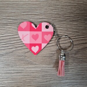 May include: A pink heart-shaped keychain with a checkered pattern and heart and X symbols. The keychain has a silver ring and a pink tassel. The background is a wooden surface.