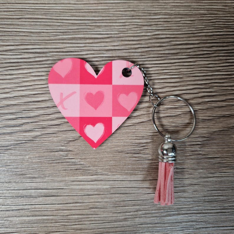 May include: A pink heart-shaped keychain with a checkered pattern and heart and X symbols. The keychain has a silver ring and a pink tassel. The background is a wooden surface.