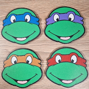 Puede incluir: Cuatro posavasos verdes y circulares con las caras de las Tortugas Ninja. Cada posavasos tiene una m&aacute;scara de diferente color: azul, morado, naranja y rojo. Los posavasos tienen contornos negros y bocas sonrientes.