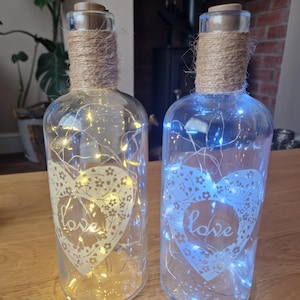 May include: Two glass bottles with cork stoppers and twine wrapped around the necks. Each bottle contains a string of white fairy lights and a white heart with the word "love" etched into it. One bottle has warm white lights and the other has blue lights.