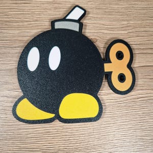 May include: A black, round cartoon bomb with white oval eyes, yellow feet, and a wind-up key. The bomb has a gray top with a white rectangle. The design is on a wooden surface.