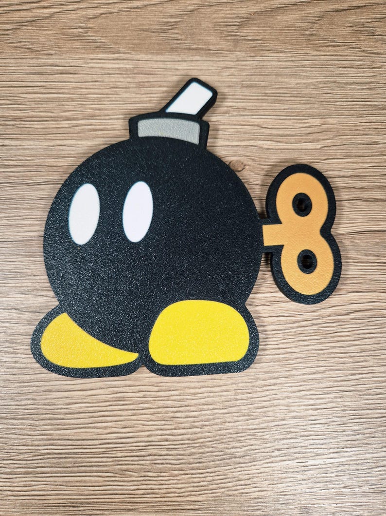 May include: A black, round cartoon bomb with white oval eyes, yellow feet, and a wind-up key. The bomb has a gray top with a white rectangle. The design is on a wooden surface.