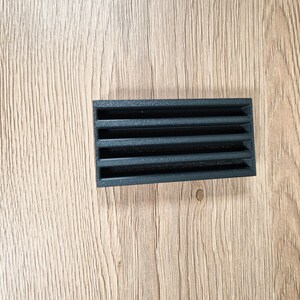 May include: A rectangular black vent cover with horizontal slats. The vent is made of a dark material and is positioned on a light brown wooden surface. The slats are evenly spaced, creating a grid-like pattern.