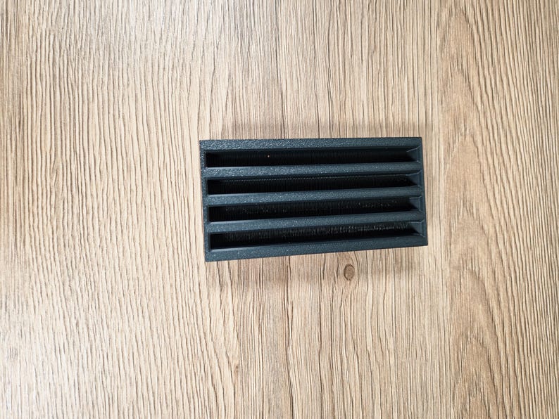May include: A rectangular black vent cover with horizontal slats. The vent is made of a dark material and is positioned on a light brown wooden surface. The slats are evenly spaced, creating a grid-like pattern.