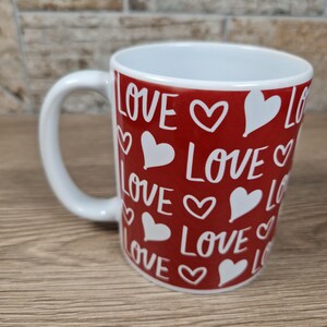 K&ouml;nnte beinhalten: Eine Keramiktasse mit wei&szlig;em Griff und roter Au&szlig;enseite. Die Tasse ist mit dem Wort "LOVE" und wei&szlig;en Herzformen verziert. Die Tasse ist f&uuml;r Getr&auml;nke konzipiert.