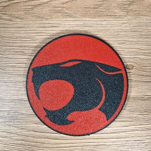 May include: A round, red and black graphic featuring a stylized cat silhouette within a circle. The cat's head is black, set against a red background. The design is centered on a wooden surface.