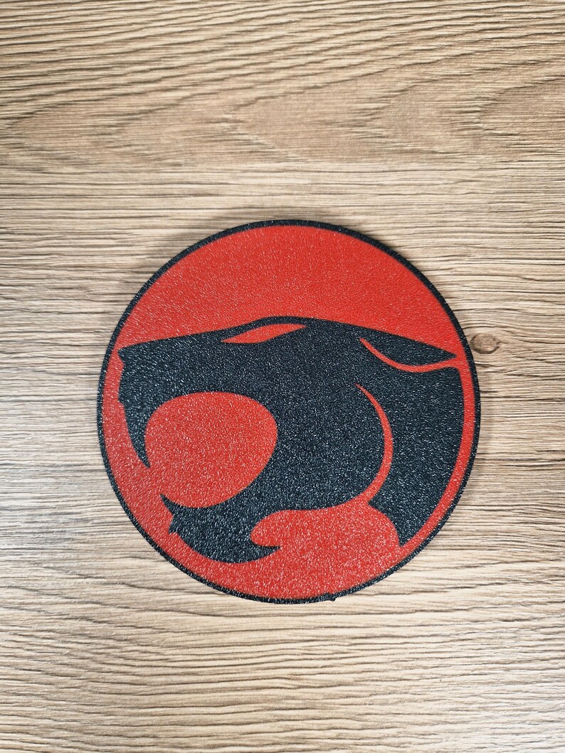 May include: A round, red and black graphic featuring a stylized cat silhouette within a circle. The cat's head is black, set against a red background. The design is centered on a wooden surface.