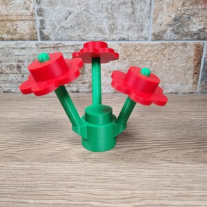 May include: A 3D-printed decorative flower arrangement. The flowers have red petals and green stems. Each flower has a green center. The base is green, and the arrangement is on a wooden surface.