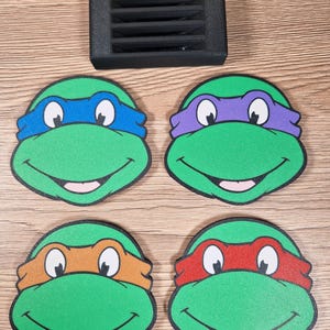 Puede incluir: Cuatro posavasos verdes de las Tortugas Ninja con cintas para la cabeza de diferentes colores: azul, morado, naranja y rojo. Un objeto rectangular negro est&aacute; encima de los posavasos. Los posavasos est&aacute;n sobre una superficie de madera.
