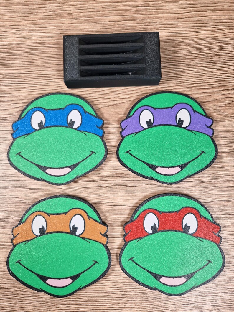 Puede incluir: Cuatro posavasos verdes de las Tortugas Ninja con cintas para la cabeza de diferentes colores: azul, morado, naranja y rojo. Un objeto rectangular negro est&aacute; encima de los posavasos. Los posavasos est&aacute;n sobre una superficie de madera.