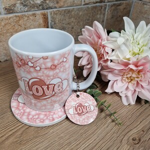 May include: A white ceramic mug, coaster, and keychain set with a pink floral and heart design. The word "Love" is featured prominently on the mug, coaster, and keychain. The mug has a white handle and is decorated with pink hearts.
