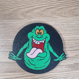 May include: A round, black coaster featuring a green cartoon character with a large mouth and tongue. The character has a smiling expression and is set against a black background. The coaster is likely made of a durable material.