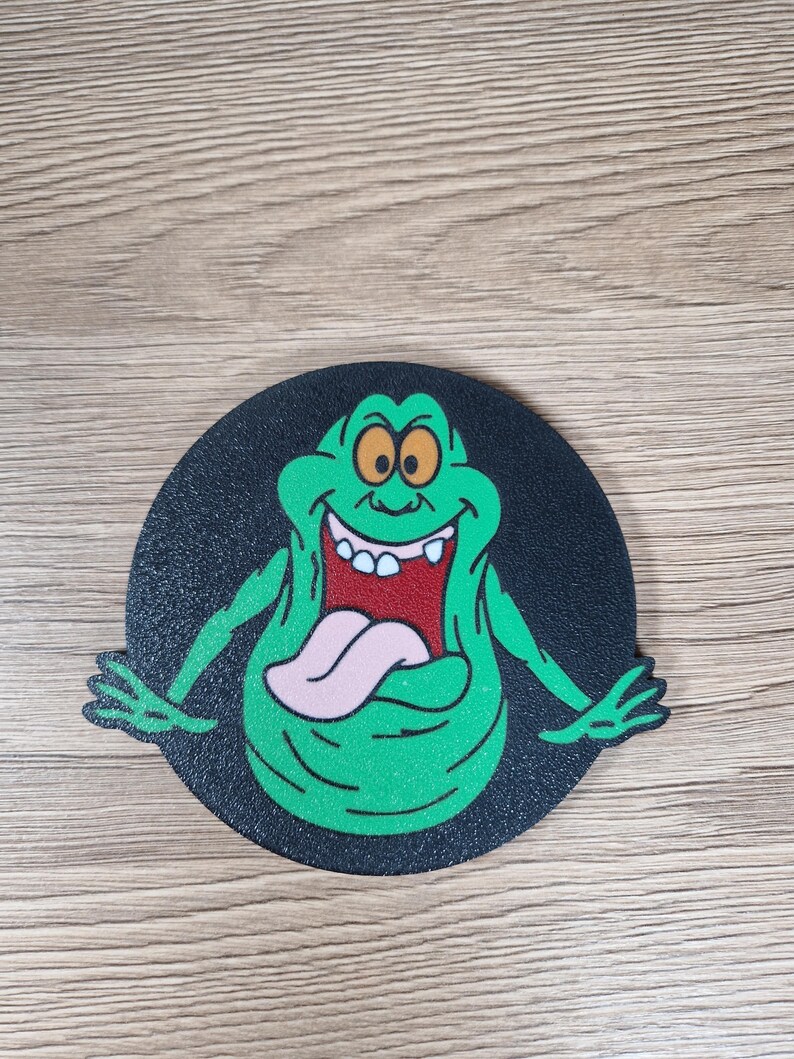 May include: A round, black coaster featuring a green cartoon character with a large mouth and tongue. The character has a smiling expression and is set against a black background. The coaster is likely made of a durable material.