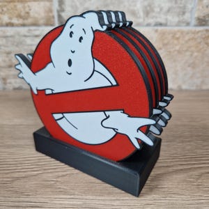 K&ouml;nnte beinhalten: Ein Set von Ghostbusters-Logo-Untersetzern. Die Untersetzer zeigen das ikonische rot-schwarze Logo mit dem wei&szlig;en Geist. Die Untersetzer sind auf einem schwarzen rechteckigen Sockel gestapelt.