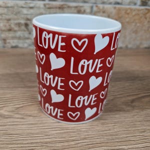 K&ouml;nnte beinhalten: Ein roter Keramikbecher mit dem Wort "LOVE" und wei&szlig;en Herzen rundherum bedruckt. Der Becher hat eine wei&szlig;e Innenseite und steht auf einer Holzoberfl&auml;che. Der Becher ist ein Artikel zum Valentinstag.
