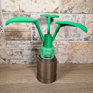 May include: A green and brown decorative item resembling a plant. The item features a barrel-shaped base, a green face, and three green leaf-like structures extending upwards. The item is made of plastic and sits on a wooden surface.