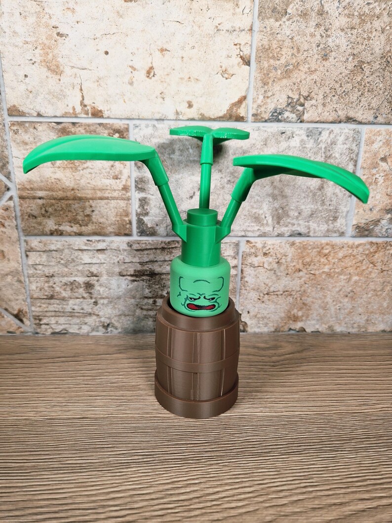 May include: A green and brown decorative item resembling a plant. The item features a barrel-shaped base, a green face, and three green leaf-like structures extending upwards. The item is made of plastic and sits on a wooden surface.