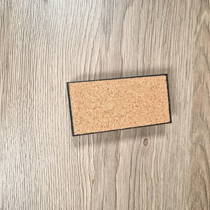 May include: A rectangular cork coaster with a black border. The coaster is made of natural cork material and is set against a light brown wooden surface. The coaster is designed to protect surfaces from heat and moisture.