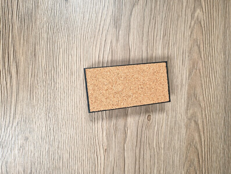 May include: A rectangular cork coaster with a black border. The coaster is made of natural cork material and is set against a light brown wooden surface. The coaster is designed to protect surfaces from heat and moisture.