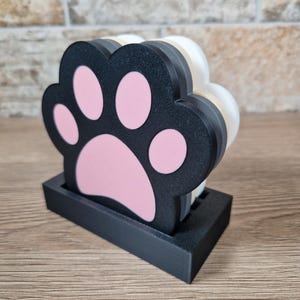 Op de afbeelding: Een set pootvormige onderzetters in een zwarte houder. De onderzetters zijn zwart met roze pootkussentjes. De houder is ook zwart. De onderzetters zijn opgestapeld en klaar voor gebruik.