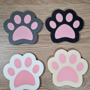 Op de afbeelding: Vier pootvormige onderzetters in verschillende kleuren. Twee zijn donkergrijs en zwart met roze pootkussentjes, en twee zijn wit en beige met roze pootkussentjes. Perfect voor dierenliefhebbers.
