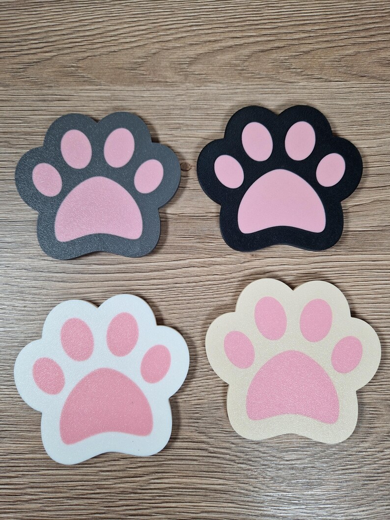 Op de afbeelding: Vier pootvormige onderzetters in verschillende kleuren. Twee zijn donkergrijs en zwart met roze pootkussentjes, en twee zijn wit en beige met roze pootkussentjes. Perfect voor dierenliefhebbers.