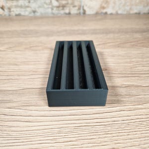 May include: A rectangular, dark gray organizer with five vertical slots. The object is made of a matte material and sits on a light wood surface. The organizer is designed to hold small items.