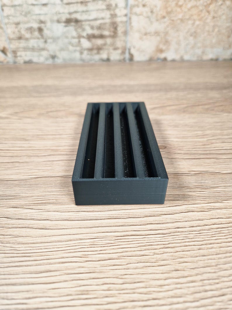 May include: A rectangular, dark gray organizer with five vertical slots. The object is made of a matte material and sits on a light wood surface. The organizer is designed to hold small items.