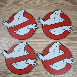 K&ouml;nnte beinhalten: Vier Ghostbusters-Logo-Untersetzer. Jeder Untersetzer zeigt eine wei&szlig;e Geisterfigur in einem roten Kreis mit einem diagonalen Strich. Die Untersetzer bestehen aus einem strukturierten Material und sind auf einer Holzoberfl&auml;che angeordnet.