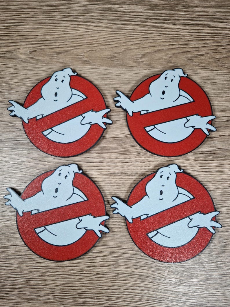 K&ouml;nnte beinhalten: Vier Ghostbusters-Logo-Untersetzer. Jeder Untersetzer zeigt eine wei&szlig;e Geisterfigur in einem roten Kreis mit einem diagonalen Strich. Die Untersetzer bestehen aus einem strukturierten Material und sind auf einer Holzoberfl&auml;che angeordnet.