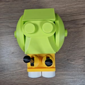 Puede incluir: Un juguete robot impreso en 3D de color verde lima y amarillo con perillas negras y pies blancos. El robot tiene una cabeza redondeada, dos ojos circulares y un cuerpo rectangular. El juguete est&aacute; hecho de pl&aacute;stico y se encuentra sobre una superficie de madera.