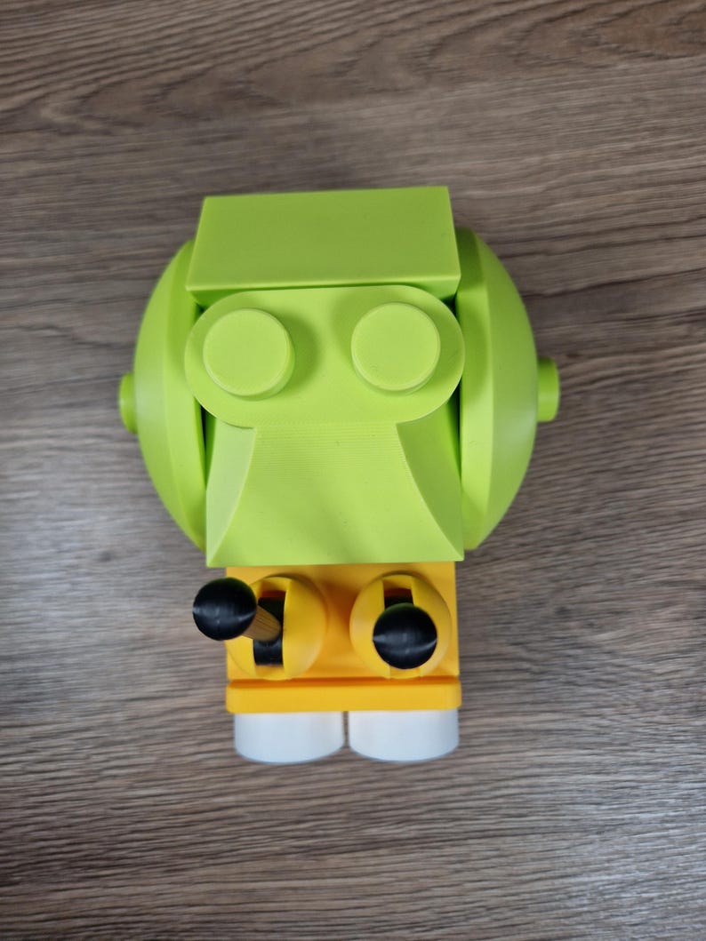Puede incluir: Un juguete robot impreso en 3D de color verde lima y amarillo con perillas negras y pies blancos. El robot tiene una cabeza redondeada, dos ojos circulares y un cuerpo rectangular. El juguete est&aacute; hecho de pl&aacute;stico y se encuentra sobre una superficie de madera.