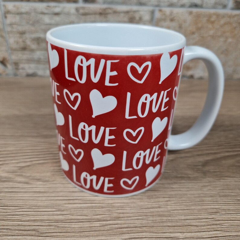 K&ouml;nnte beinhalten: Eine wei&szlig;e Keramiktasse mit rotem Hintergrund. Die Tasse ist mit dem Wort "LOVE" und wei&szlig;en Herzformen verziert. Die Tasse hat einen wei&szlig;en Henkel und einen wei&szlig;en Innenraum.