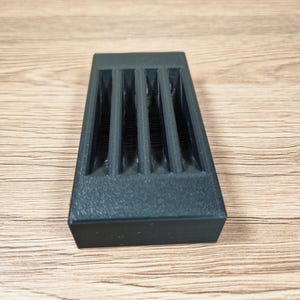 Puede incluir: Un objeto rectangular gris oscuro con cuatro ranuras verticales. El objeto tiene una superficie texturizada y est&aacute; sobre una superficie de madera.