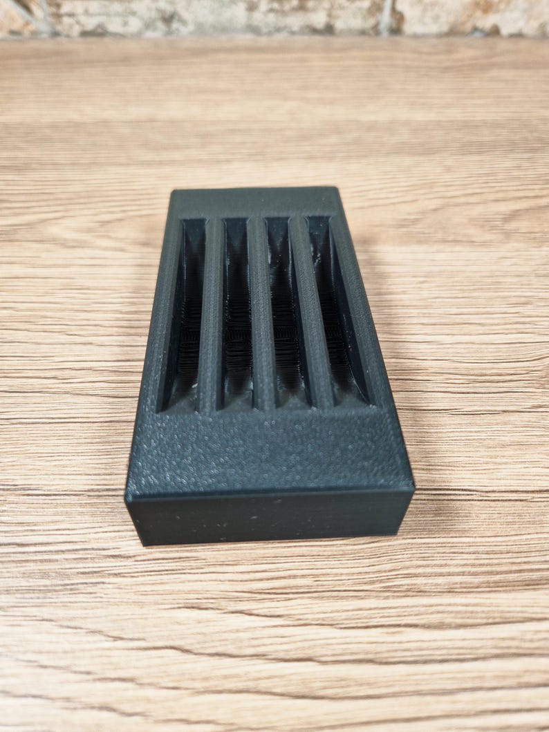 Puede incluir: Un objeto rectangular gris oscuro con cuatro ranuras verticales. El objeto tiene una superficie texturizada y est&aacute; sobre una superficie de madera.