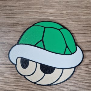 K&ouml;nnte beinhalten: Eine gr&uuml;ne und schwarze Koopa Troopa-Schale mit wei&szlig;em Rand und beigen Augen. Die Schale hat eine strukturierte Oberfl&auml;che und ist eine erkennbare Figur aus der Super Mario-Videospielserie.