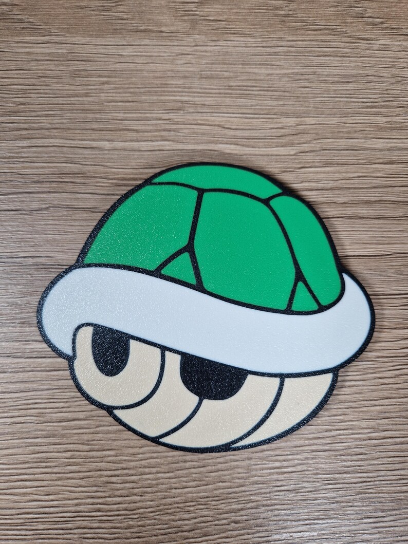 K&ouml;nnte beinhalten: Eine gr&uuml;ne und schwarze Koopa Troopa-Schale mit wei&szlig;em Rand und beigen Augen. Die Schale hat eine strukturierte Oberfl&auml;che und ist eine erkennbare Figur aus der Super Mario-Videospielserie.