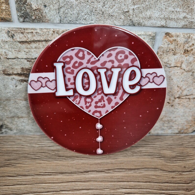 May include: A round, red decorative item with the word "LOVE" in white block letters. A heart-shaped design with a leopard print pattern is in the center. The item has a white border with pink hearts and small white hearts hanging below.