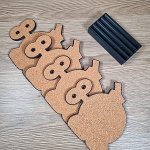 May include: Set of four cork coasters in a pig shape with black outlines and a black holder. The coasters are light brown with a textured surface. The holder has four slots to store the coasters. The coasters are approximately 4 inches wide.