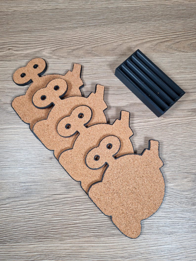 May include: Set of four cork coasters in a pig shape with black outlines and a black holder. The coasters are light brown with a textured surface. The holder has four slots to store the coasters. The coasters are approximately 4 inches wide.
