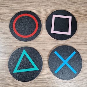 K&ouml;nnte beinhalten: Ein Set aus vier runden, schwarzen Untersetzern, die jeweils mit einem farbigen geometrischen Symbol versehen sind: ein roter Kreis, ein rosa Quadrat, ein t&uuml;rkisfarbenes Dreieck und ein blaues Kreuz. Die Untersetzer haben eine texturierte Oberfl&auml;che.